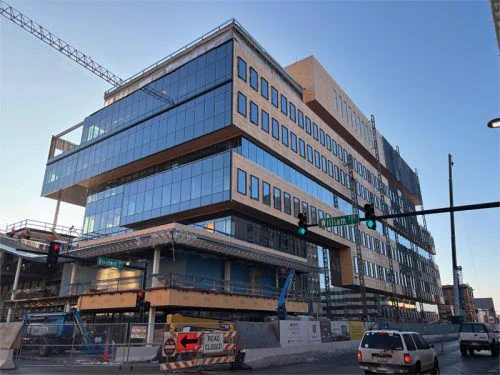 Modern glass building under construction at a city intersection. Cranes, fencing, and a "Road Closed" sign indicate active development work.