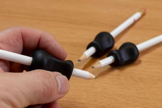 A hand holds a pencil with a black ergonomic grip. Two more pencils with grips are in the background.
