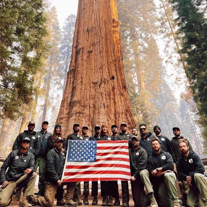 Group of hotshots posing with an American flag in front of a massive sequoia tree in a forest.