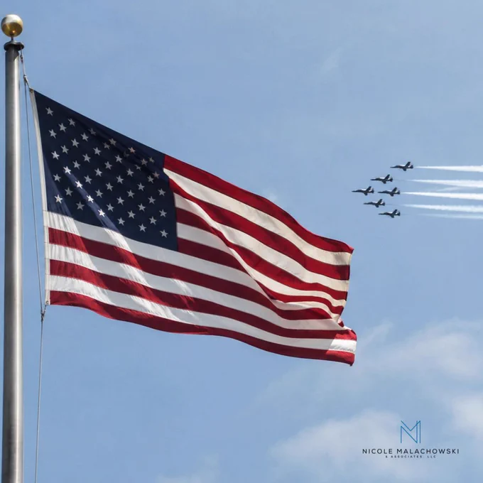 American flag waving on a flagpole with military jets flying in formation across a clear sky.