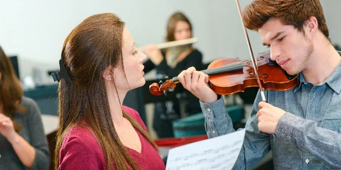 A violinist plays while a person in a red top holds sheet music during a music lesson in a studio.
