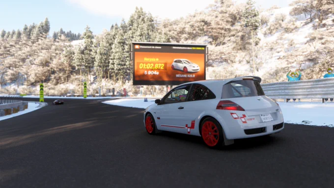 A white race car with red accents is on a snowy track near a curve. In the background, there's a large digital billboard displaying a racing time of 1:02.872 and an image of a Renault Mégane R.S. The surrounding area is covered with snow, and there are trees in the background.