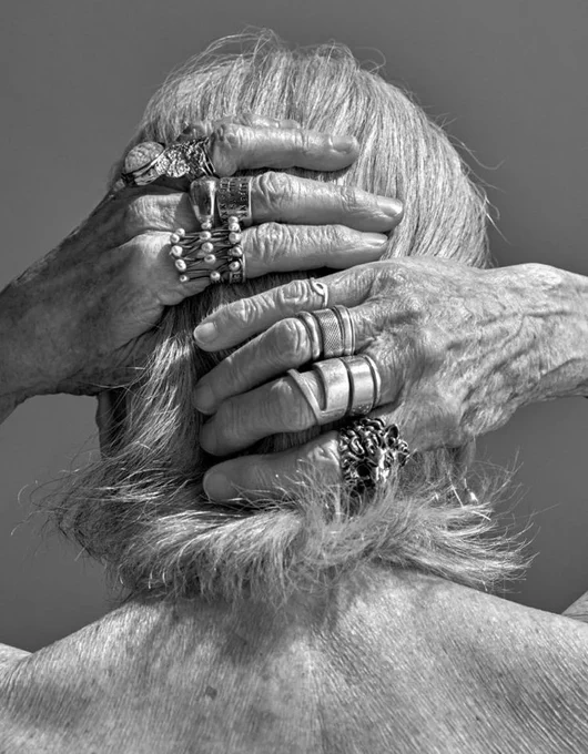 Black and white high-contrast photo of hands laced behind a grey haired head, adorned with several chunky silver rings.
