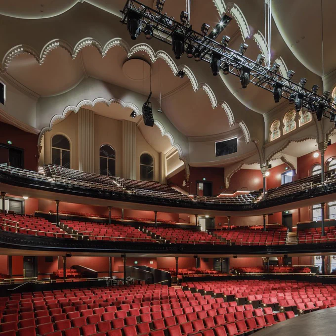 A large theatre auditorium with rows of red seats facing a stage in the lower right foreground. Multiple balcony levels wrap around the room, with dark railings, gold accents, arched windows, stained glass panels and red walls. The ceiling has ornate scalloped arches and curved plasterwork. A long black lighting rig with stage lights hangs above the seating area.
