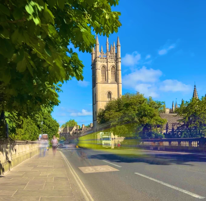 Image shows a sunny day in Oxford with the iconic Magdalen Tower visible above lush trees. Blurry figures can be seen walking along Magdalen Bridge.