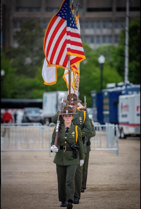 A group of uniformed individuals is participating in a procession, with one person holding the American flag. The setting appears to be outdoors with a fenced area and large structures in the background.
