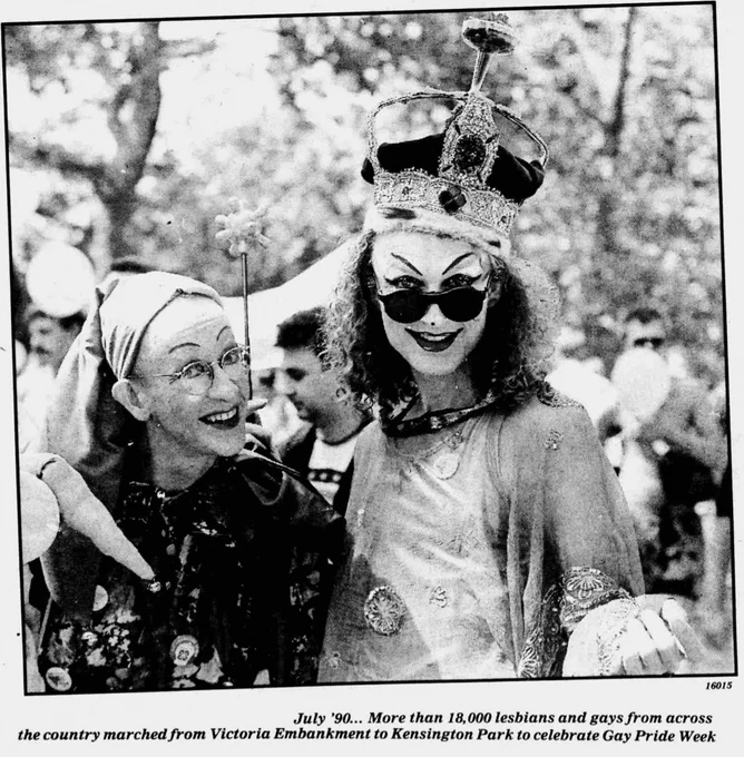 A joyful scene from Gay Pride Week in July 1990, featuring two individuals in vibrant and festive attire. One is adorned with an ornate crown and decorative accessories. They stand amidst a leafy outdoor setting, celebrating with many others who marched from Victoria Embankment to Kensington Park.