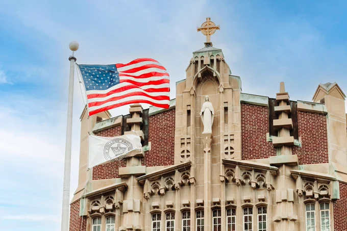 A large Catholic church facade with intricate stonework, featuring a statue and a cross at its pinnacle. An American flag and a white flag with a crest are flying in front, set against a blue sky with clouds.