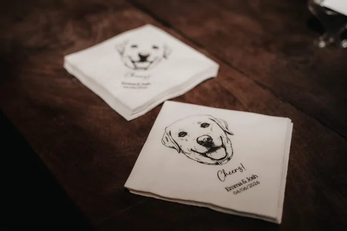 White napkins are on a wooden table. There is a drawing a dog on them with "cheers!" written.