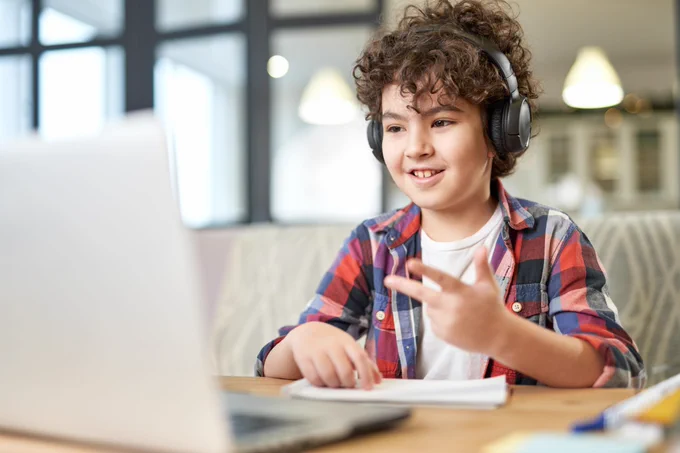 Child wearing headphones and a plaid shirt engaged in an online learning session with a laptop and notebook on the table.
