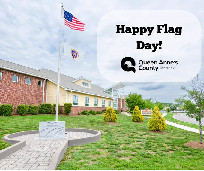 American and Maryland state flags flying on a pole outside a brick and beige building under a cloudy sky.