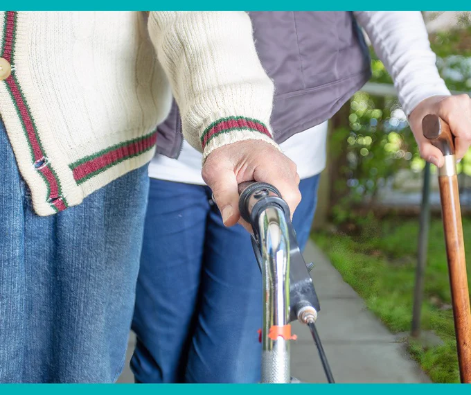 Close-up of two elderly individuals walking outdoors, one using a walker and the other a wooden cane for support.