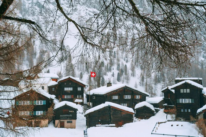 Picturesque scenery of modern village cottages with Switzerland flag located on forested snowy hillside on cold winter day