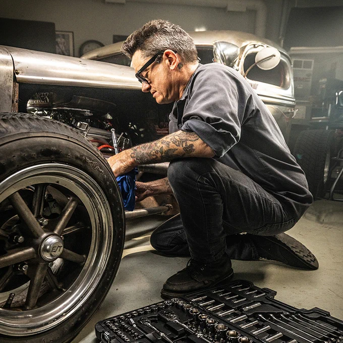Man with tattooed arm working on a vintage car engine in a garage, surrounded by tools on the floor.