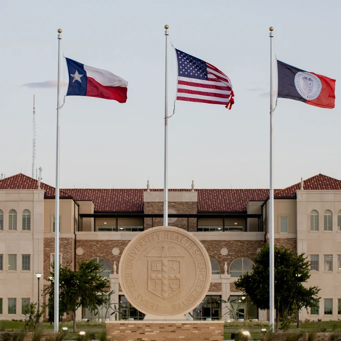 Three flags flying behind the TTUHSC seal with a building in the background.
