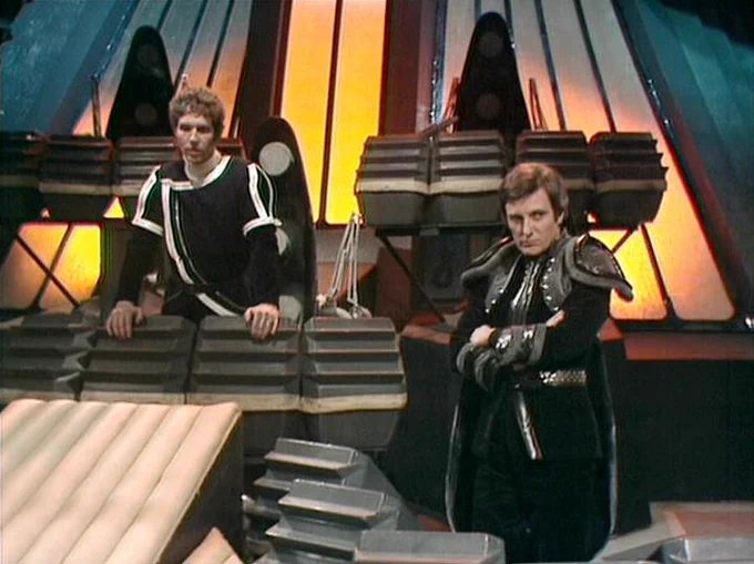 GPT 5.4 Nano describes the image as: "The image shows a tense scene inside a futuristic set from the BBC science-fiction series Blake’s 7. Two men in dark, tailored uniforms stand amid stepped metal platforms and large, curved wall panels glowing with warm orange light. Steven Pacey appears seated or leaning on a blocky control surface on the left, watching toward the action. Paul Darrow stands on the right, holding a device or weapon-like instrument close to his chest as if preparing to use it. The background is dominated by industrial grey structure, speakers or vented fixtures, and dramatic lighting that suggests a high-tech command or control area."
The script search url for this tweet is
https://blake.torpidity.net/s/312/263