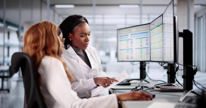 Two nurse scientists in lab coats sit at a desk with multiple computer monitors displaying data.