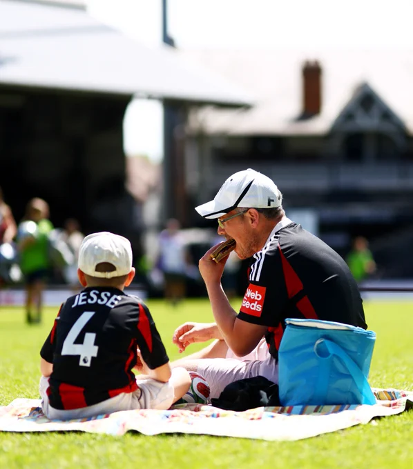 Two Fulham fans eat sandwiches on the pitch at Craven Cottage, with the cottage pictured in the background.