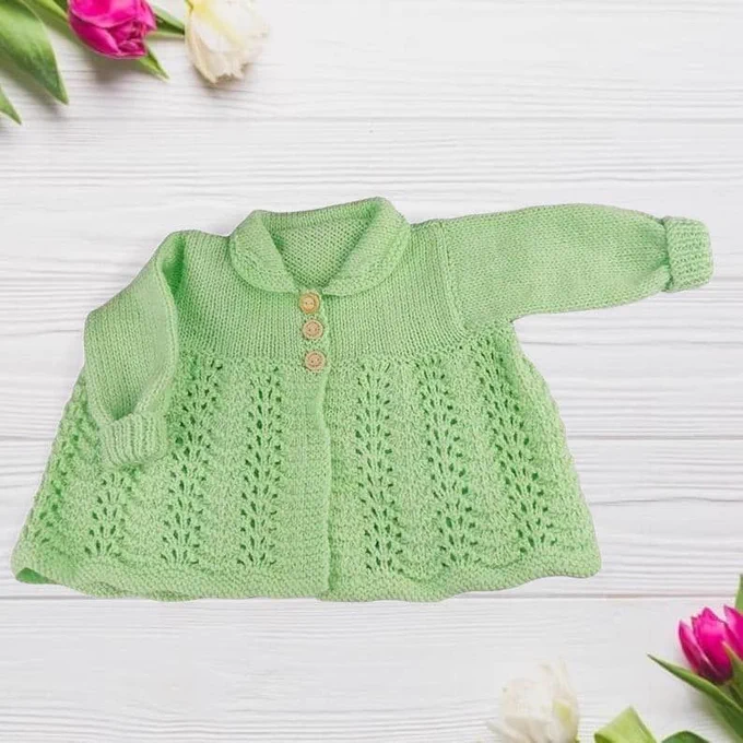 A mint green knit baby cardigan with a rounded collar and three buttons lies on a white wood surface. It features a decorative lace-patterned lower half. Pink and white tulips are tucked into the upper-left and lower-right corners.