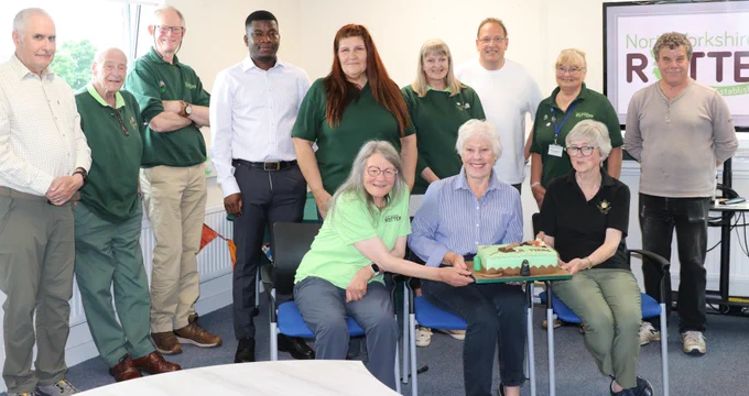A group of people, some in green shirts, gathered around a cake. A screen in the background shows "North Yorkshire ROTTER".