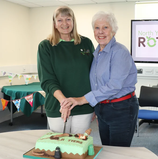 Two women cutting a cake decorated with a worm and gardening tools, celebrating "North Yorkshire Rotters".