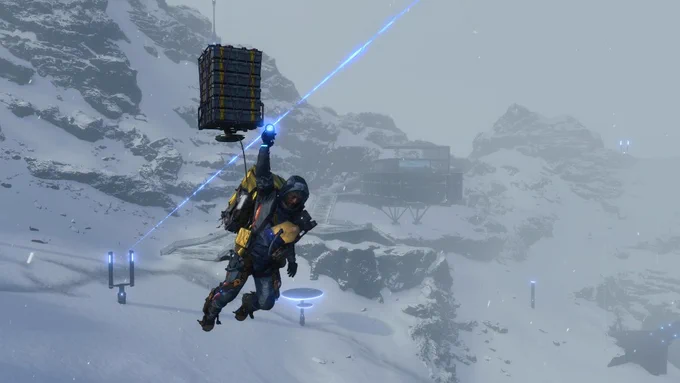 Sam Bridges aus Death Stranding Directors Cut nutzt eine Seilrutsche in den verschneiten Bergen. Er gleitet mit schwerer Fracht auf dem Rücken durch einen Schneesturm vor einer Basisstation.