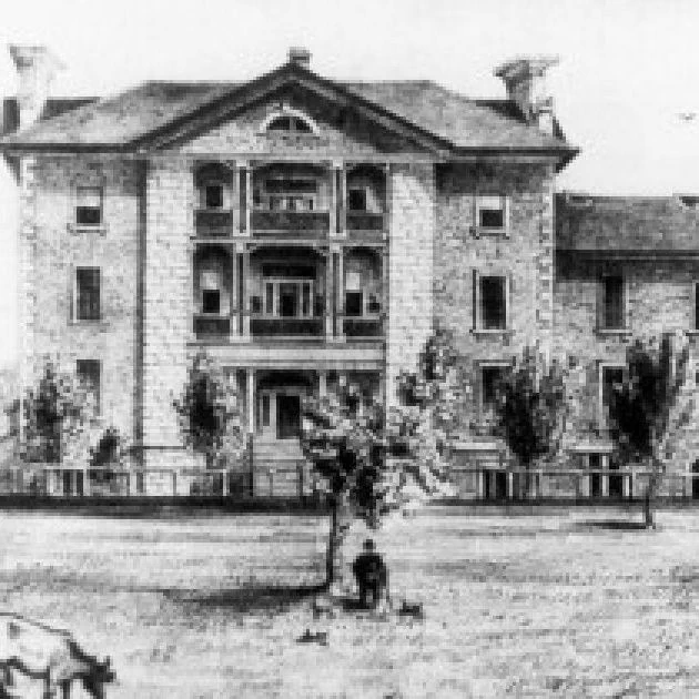 Black-and-white historical image of a large stone institutional building with multiple storeys, balconies, tall windows, and a fenced lawn in front. A few trees, animals, and small figures appear on the grounds, giving the scene a rural or campus-like setting.
