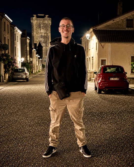 A smiling man stands confidently on a quiet street at night, wearing a black hoodie and khaki pants, with a historic tower in the background.