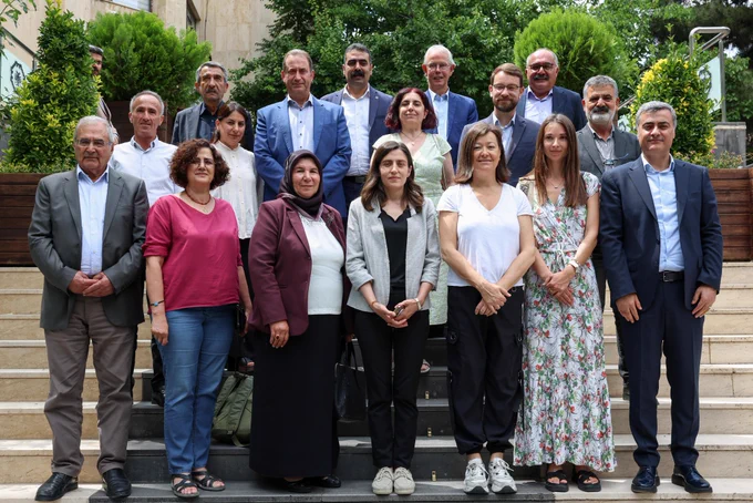 Görsel (Dış mekân grup fotoğrafı)

* Yaklaşık 18–20 kişilik bir grup, bir binanın girişindeki merdivenlerde poz vermiş.
* Arka planda yeşil ağaçlar, çalılar ve taş kaplamalı bir bina bulunuyor.
* Katılımcıların çoğu iş veya yarı-resmî kıyafetler giymiş:
    * Erkeklerin büyük bölümü ceket, gömlek ve takım elbise tercih etmiş.
    * Kadınlar arasında ceket, bluz, uzun etek ve elbise giyenler var.
* Grup üç sıra halinde düzenlenmiş; öndekiler merdivenin alt kısmında, diğerleri daha yukarı basamaklarda duruyor.
* Fotoğrafın genel havası kurumsal, profesyonel ve resmî bir etkinliği çağrıştırıyor.