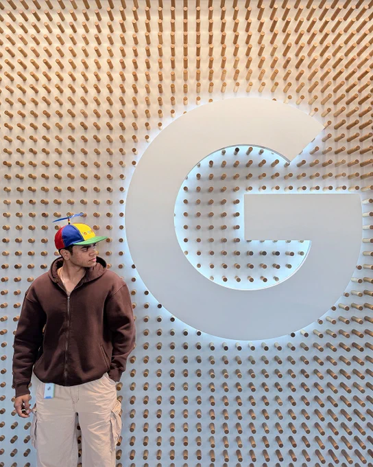 Smit wearing a brown jacket and a Noogler hatting looking at a white G logo on a wall. 
