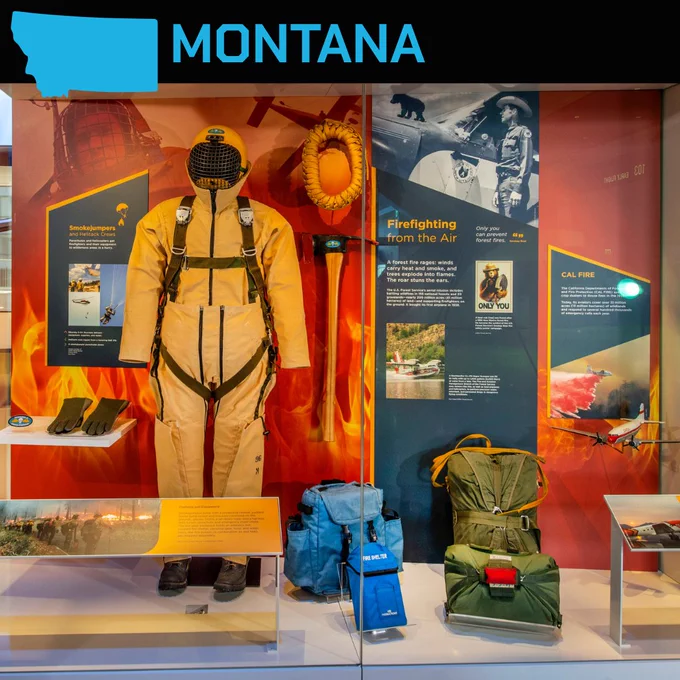 A display featuring fire-fighting equipment and informational panels. The exhibit includes a firefighting suit, helmet, gloves, and various tools. Panels detail firefighting methods with images of aircraft used in operations. The top has a "Montana" label alongside a state outline.