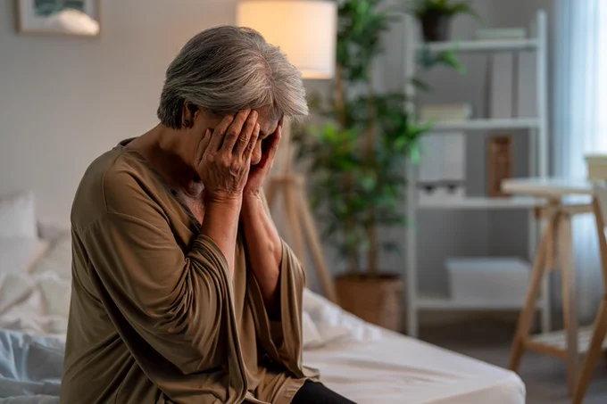 Elderly person sitting on a bed with hands covering their face, appearing distressed in a softly lit bedroom.