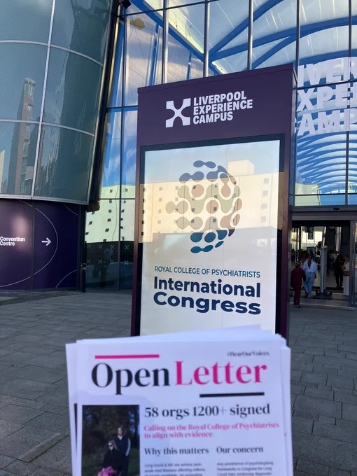 A photograph taken outdoors showing a hand holding a printed pamphlet in the foreground, with a large digital sign and a modern building in the background. The blurred pamphlet in the lower frame displays the heading "Open Letter" in a bold serif font, accompanied by the text "58 orgs 1200  signed" and "#HearOurVoices". In the center background stands a tall, purple digital monolith sign under the branding "LIVERPOOL EXPERIENCE CAMPUS". The screen displays a colorful, abstract circular mosaic logo above the text "ROYAL COLLEGE OF PSYCHIATRISTS International Congress". Behind the sign is the glass facade of a modern convention centre reflecting the blue sky and nearby buildings, with an entrance on the right where people are walking through.