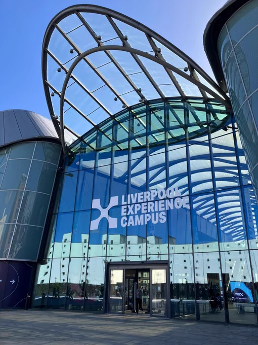 An exterior, low-angle shot of a modern convention center entrance featuring a grand, multi-story glass facade and an arched steel canopy roof under a clear blue sky. Large white lettering across the glass front reads "LIVERPOOL EXPERIENCE CAMPUS" next to a stylized "X" logo. The reflective glass panels mirror nearby white buildings and a bright, sunny sky. A person is visible walking through the glass sliding doors at the base of the entrance.