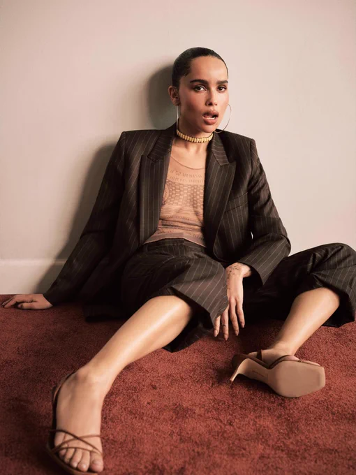 Zoë Kravitz posa seduta sul pavimento contro una parete grigio chiaro. Indossa un elegante completo gessato scuro composto da una giacca oversize lasciata aperta e pantaloni coordinati. Sotto la giacca porta una maglia semitrasparente color carne con ricami geometrici sul petto. Il look è completato da grandi orecchini a cerchio dorati, una collana a choker rigida, sandali marroni con tacco alto e punta squadrata, e una moquette rossa sullo sfondo.