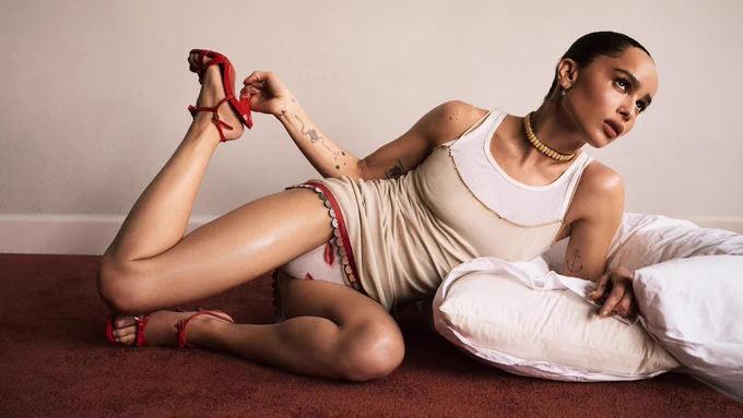 Zoë Kravitz è sdraiata su un tappeto rosso scuro, appoggiata a un cuscino bianco. Indossa una canottiera beige sopra una bianca, slip chiari decorati e sandali rossi con tacco alto e cinturino alla caviglia. Con la mano destra tiene il cinturino della scarpa del piede sollevato. Ha i capelli raccolti, una collana a maglie dorate e diversi tatuaggi visibili sulle braccia. Lo sfondo è una parete minimalista color crema.