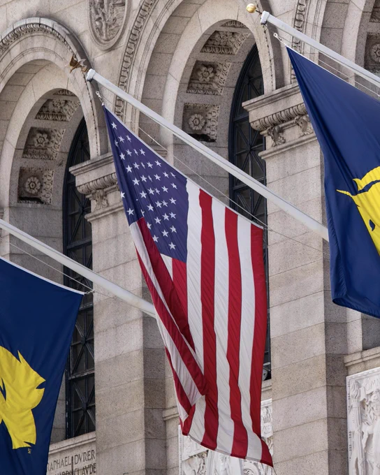 Photo of an American flag (with a B.A.A. flag on each side) hanging from the Boston Public Library in Copley Square