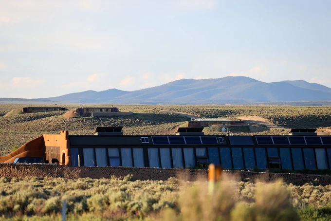 A sustainable building with solar panels on the roof stands in a vast desert landscape near Taos, New Mexico. The surrounding area features sparse vegetation and distant mountains under a clear sky.