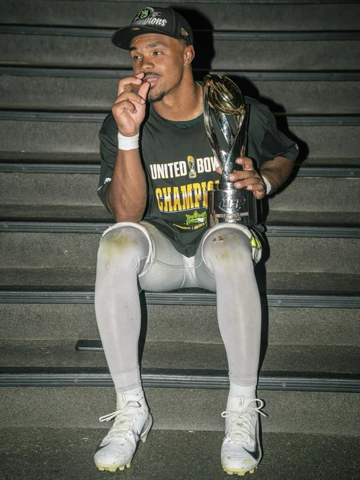 A football player celebrates on a staircase, holding a trophy and wearing a championship shirt and cap, exuding pride and victory.