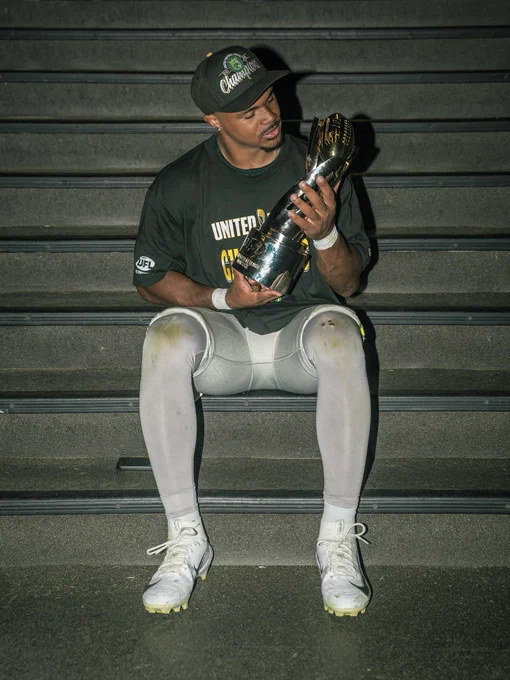 An athlete sits on steps holding a trophy, wearing a 'Champions' cap and shirt, looking at the trophy with pride.