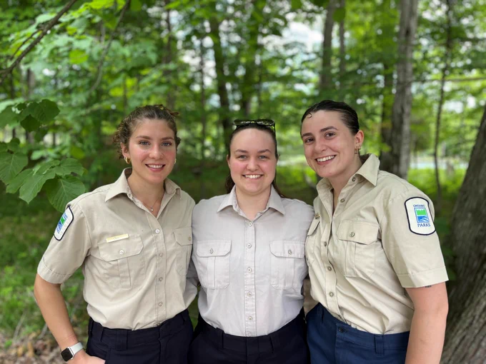 Three people in Ontario Park uniforms, standing side by side and smiling. In the background is a green forest.