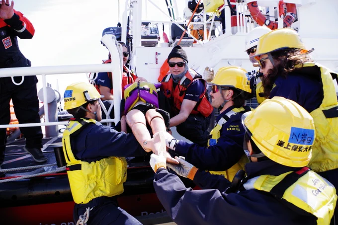 Rescue team wearing helmets and safety gear assisting in lifting a person onto a boat near the water, with hoisting equipment visible.