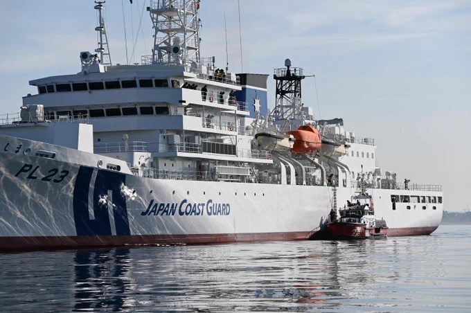 Large Japan Coast Guard white vessel sailing on calm water, accompanied by a small boat nearby, with visible structures and equipment on deck.