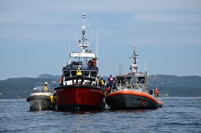 Three rescue vessels sit side by side on calm water, with personnel visible on board and a shoreline in the background.