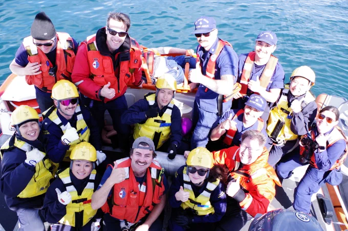 Group of people wearing life jackets and helmets gathered in a boat, looking toward the camera on calm water.