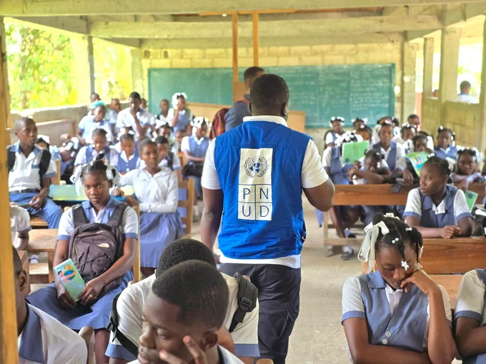 A man wearing a blue PNUD vest stands in front of a classroom filled with attentive students in school uniforms.