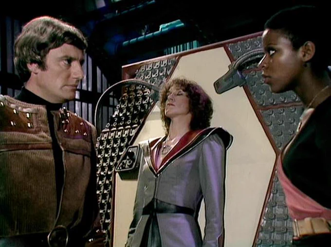GPT 5.4 describes the image as: "A tense confrontation unfolds in a futuristic interior from the BBC series Blake’s 7. At left, Paul Darrow appears as Avon, his guarded expression and rigid posture suggesting calculation and mistrust. Center frame, Jan Chappell stands within a doorway-like structure as Cally, eyes closed and face composed, giving the moment a ceremonial or psychologically charged feel. At right, Josette Simon appears as Dayna, facing Avon with clear intensity, her stance alert and confrontational. The metallic walls, geometric panels, and mounted device emphasize the show’s distinctive late-1970s science-fiction production design. Their costumes—structured tunics, bold trims, and practical belts—place the scene firmly aboard a technologically advanced setting, while the actors’ body language conveys the interpersonal strain and moral complexity that define Blake’s 7."
The script search url for this tweet is
https://blake.torpidity.net/s/310/279