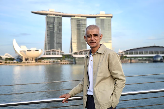 Mayor of London Sadiq Khan stands by a waterfront railing with a large modern hotel complex (Marina Bay Sands) in the background. The structure has three tall towers topped by a long, boat-shaped roof deck. 