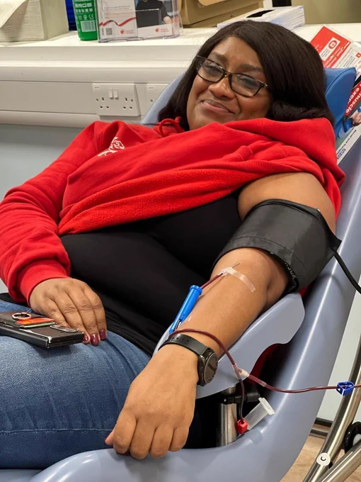 Bell Ribeiro-Addy MP reclines in a padded medical chair inside a clinical setting. A blood donation or medical tube is inserted into the person’s inner elbow, with dark red tubing leading to equipment off-frame. A black blood pressure cuff is wrapped around the upper arm. The person is wearing a red hoodie, a black top, and blue jeans, with a smartwatch on the wrist. One hand rests on the lap holding a small wallet or card holder. In the background, there are shelves with medical supplies and informational leaflets, as well as visible wall outlets and equipment.