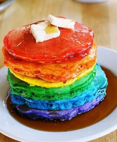 A stack of rainbow-coloured pancakes on a white plate, arranged in Pride flag colours from purple and blue at the bottom to green, yellow, orange, and red on top. The pancakes are covered in syrup, with two small pats of butter melting on the top pancake.